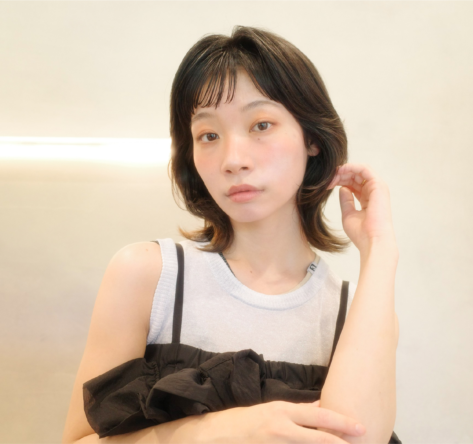 明るい背景の前で、肩を出した白いトップスと黒いワンピースを重ね着した女性が、手で髪に触れながらカメラを見つめているポートレート。