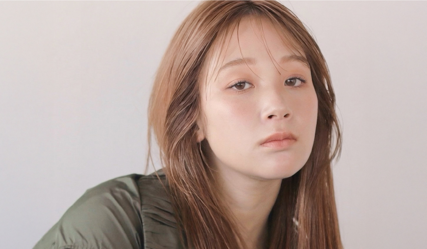明るい背景の前で、カーキ色のアウターを着たロングヘアの女性が、落ち着いた表情でカメラを見つめているポートレート。