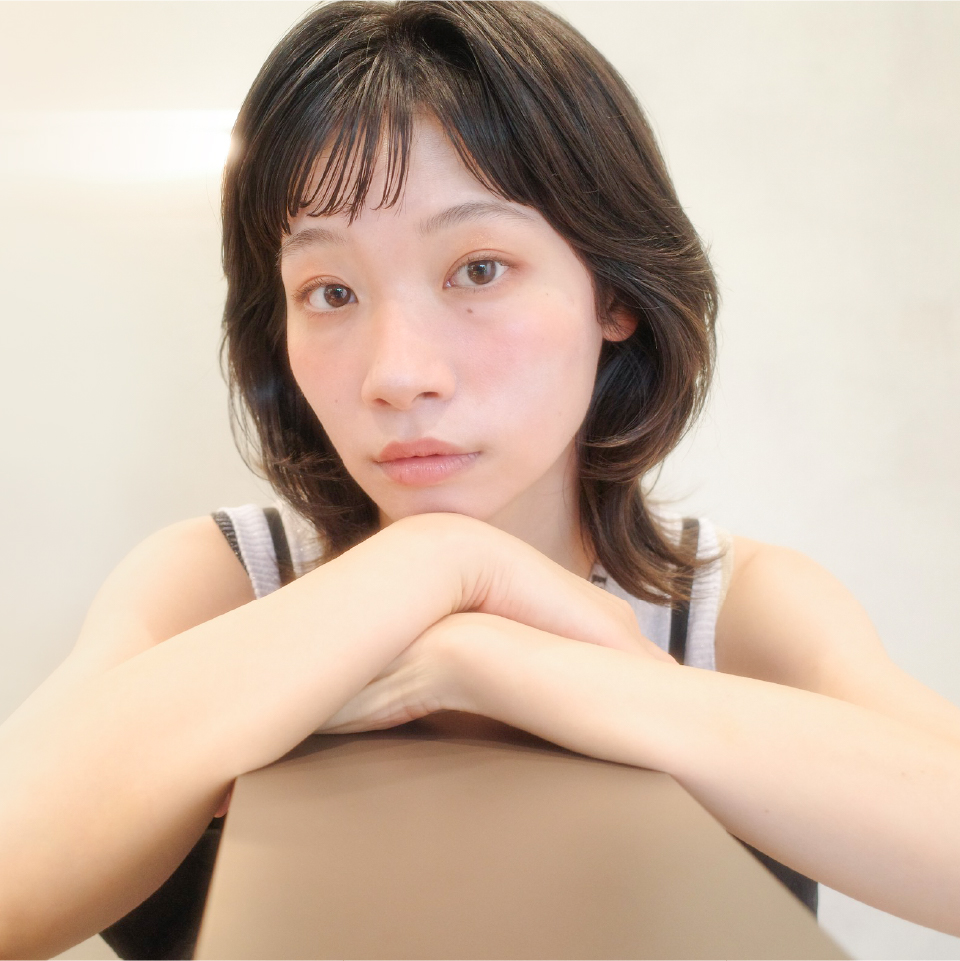 明るい室内で、女性が腕をテーブルに置き、穏やかな表情でカメラを見つめているアップのポートレート。